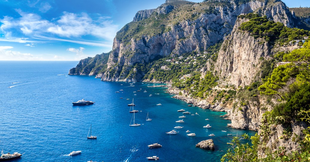 a shining fleets of superyachts off of the coast of capri with their guests enjoying the hot Mediterranean sun as they cruise from port to port on their Italy yacht charter adventure 