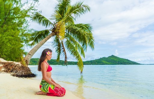 local woman knelt on the sandy beach underneath a palm tree looking out to the ocean in the French Polynesia 